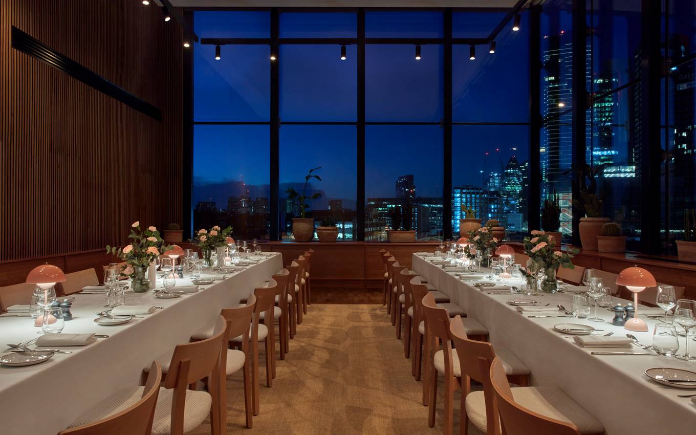 A spacious dining room with long tables set for a dinner, featuring flowers and modern lamps, with night time London views.