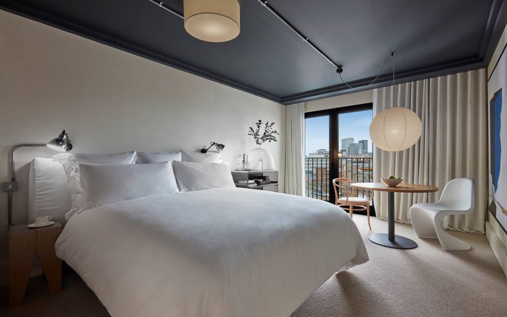 Modern hotel bedroom at One Hundred Shoreditch with a white bed, small table and a balcony view of the London skyline.