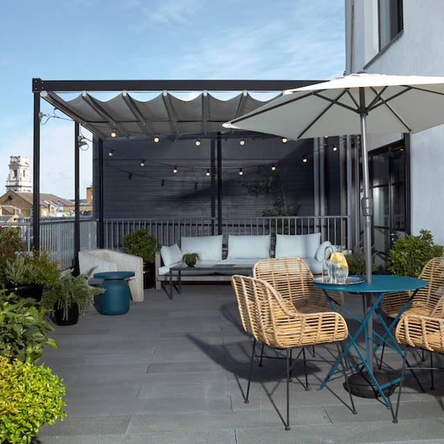 Cosy rooftop lounge area with rattan chairs at One Hundred Shoreditch with a blue table, string lights and London views.