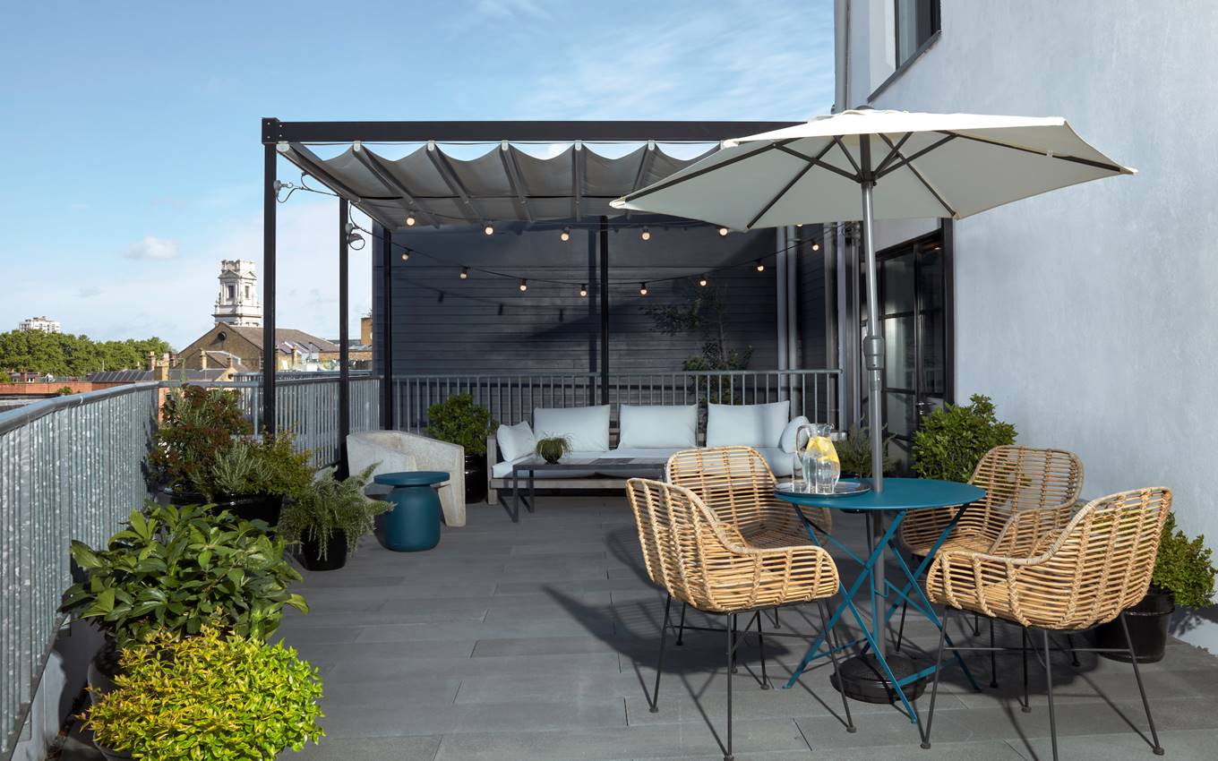 Cosy rooftop lounge area with rattan chairs at One Hundred Shoreditch with a blue table, string lights and London views.