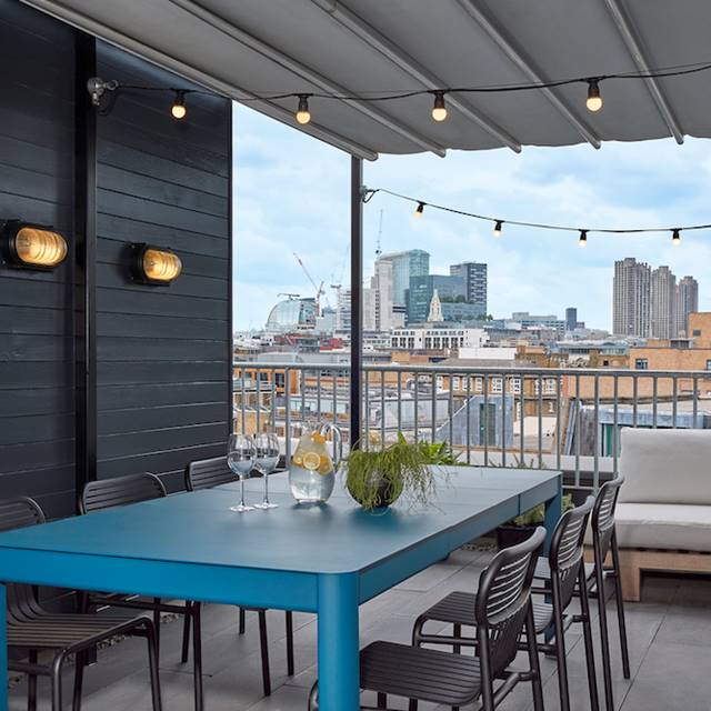 A rooftop terrace at One Hundred Shoreditch with a blue table set with glasses, offering London views under string lights.