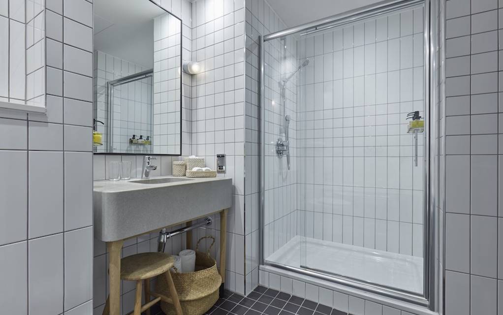 A sleek bathroom with white tiled walls, a glass-enclosed shower, a modern sink, and a wooden stool with a woven basket.