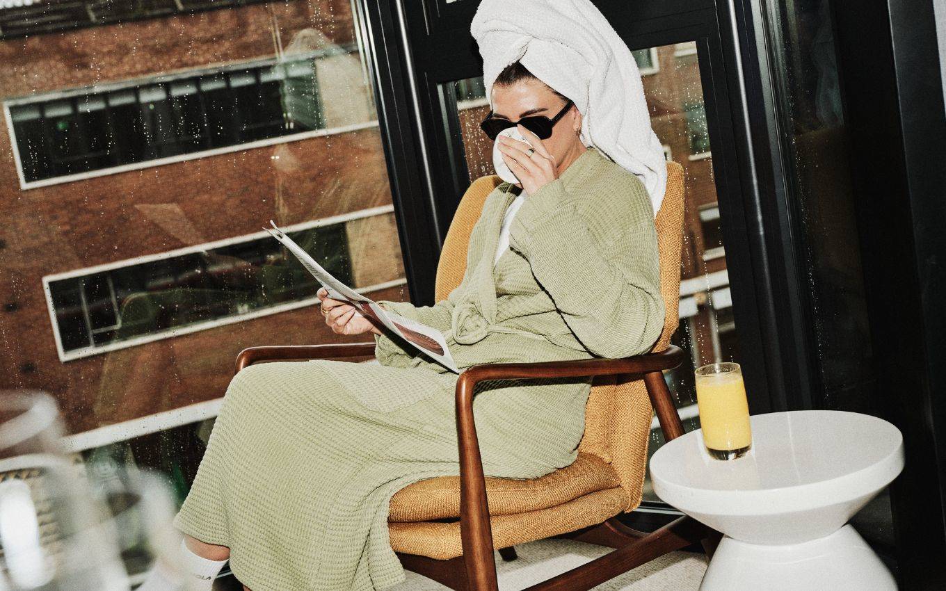 A woman in a robe and sunglasses sips a drink while reading by a rainy window at a hotel room in London.