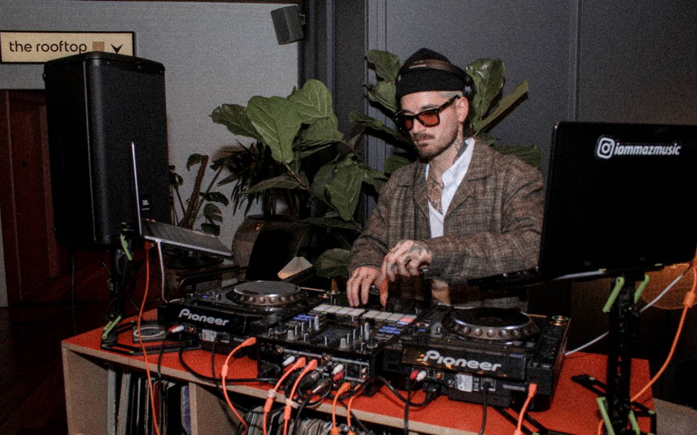 A DJ in sunglasses and a plaid jacket performing at a London bar, with Pioneer equipment and plants in the background.