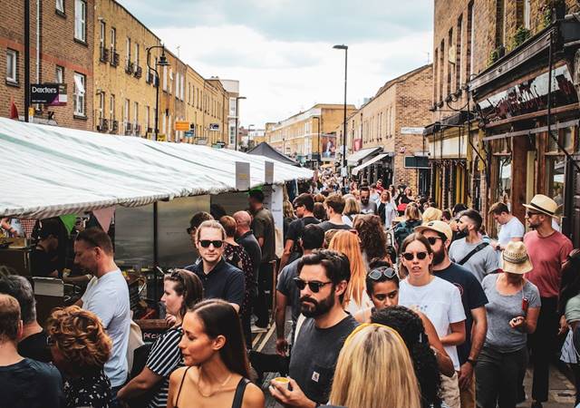 Crowded Broadway Market in London with people shopping and interacting near vendor stalls under striped canopies.