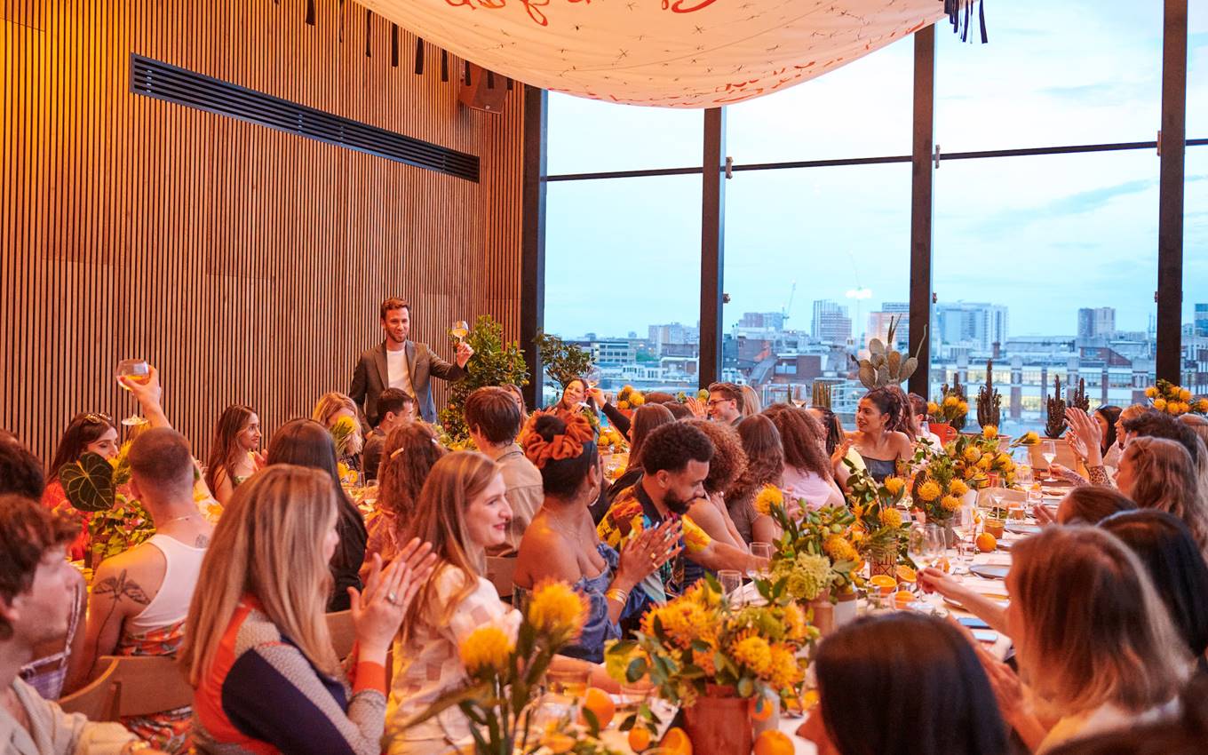 An event space in London with long tables decorated with yellow flowers, filled with guests enjoying a city view.