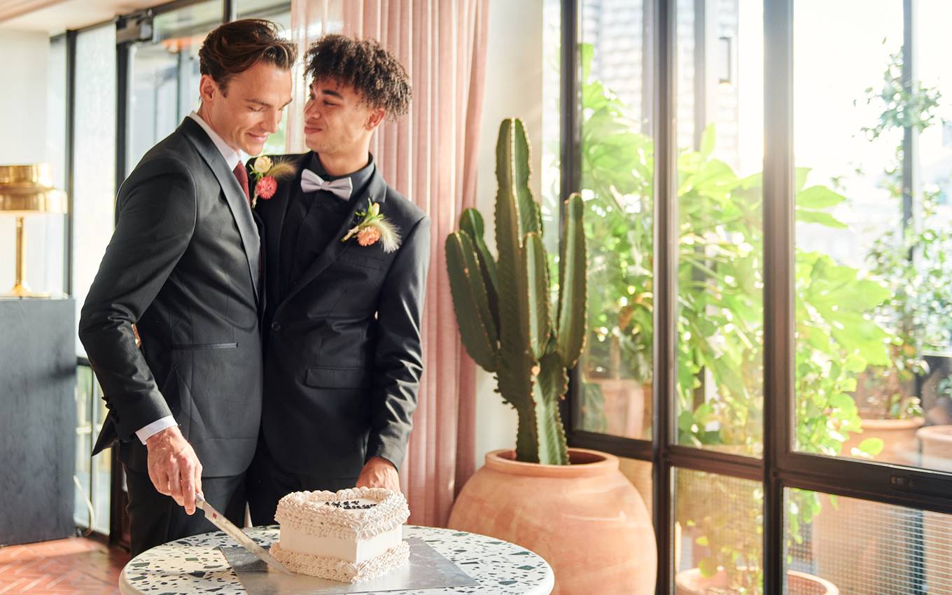 Two men in suits sharing a wedding moment while cutting a cake, standing beside large potted plants and glass windows.