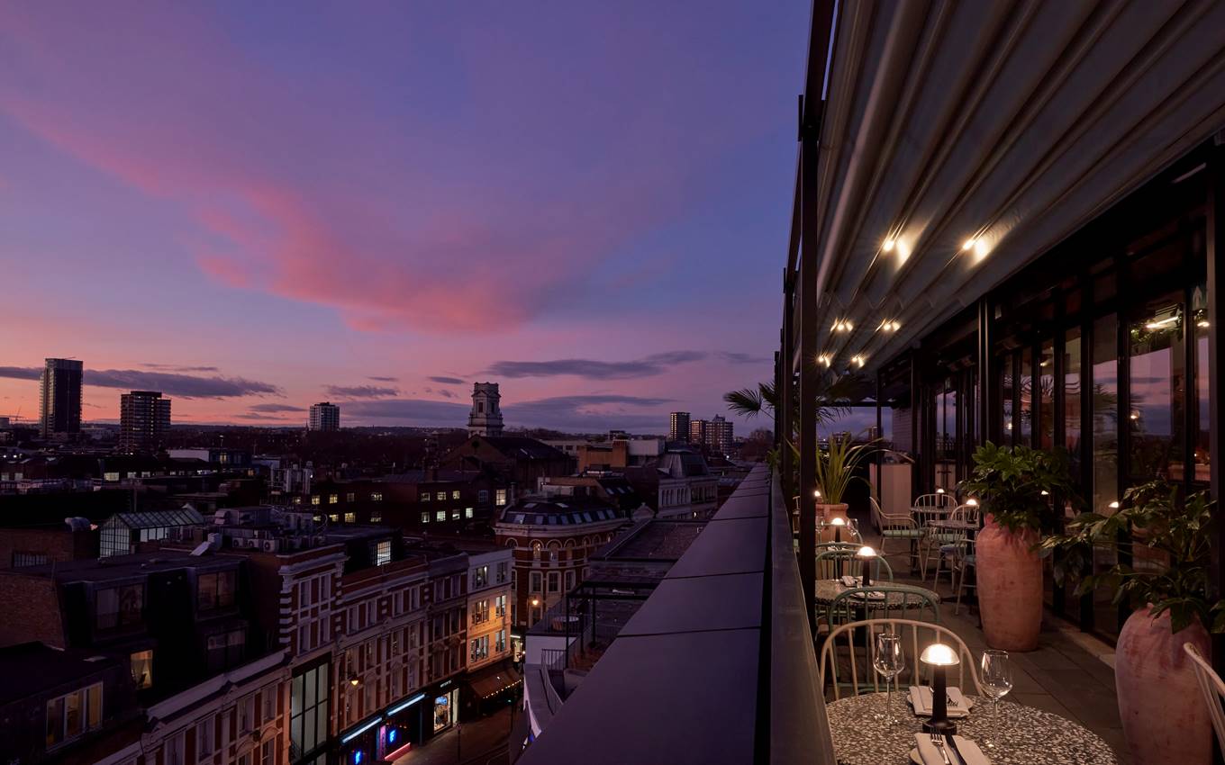 Sunset view from a rooftop terrace, overlooking the city buildings of London and the pink-purple sky, with outdoor seating.