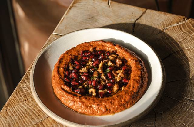 Plate with a rustic dip topped with pomegranate seeds and walnuts, on a wooden table in natural light at a London restaurant.