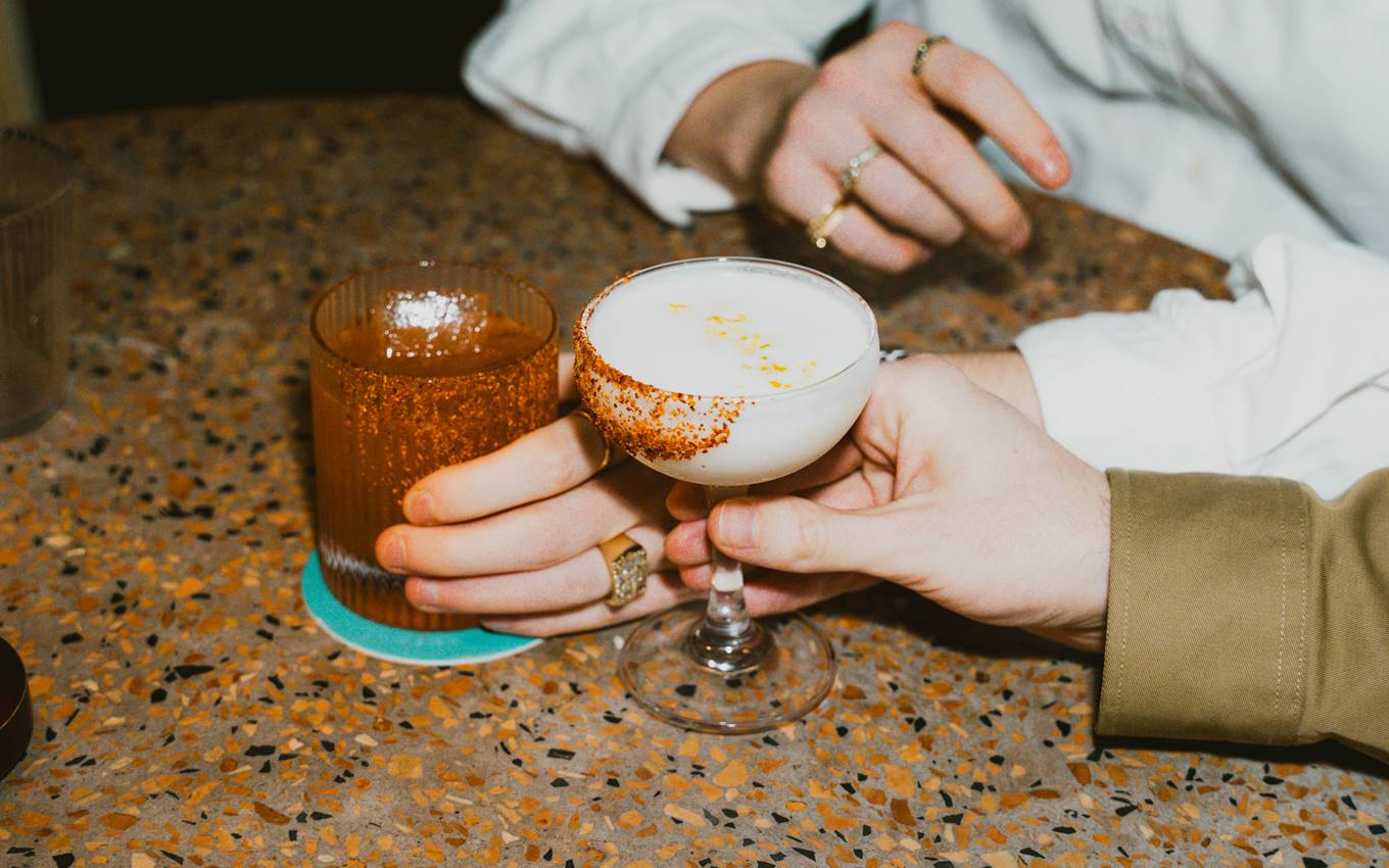 Two hands holding cocktails—one in a coupe glass with a chili rim, the other in a textured tumbler—on a speckled table.