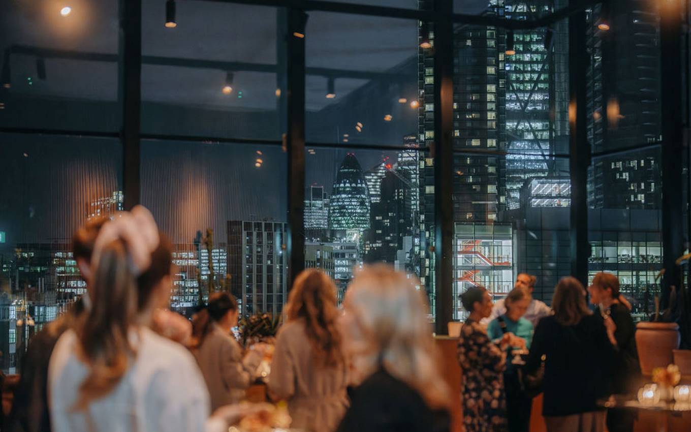 A bustling evening event with guests socializing in a lounge with large windows framing London’s cityscape at night.