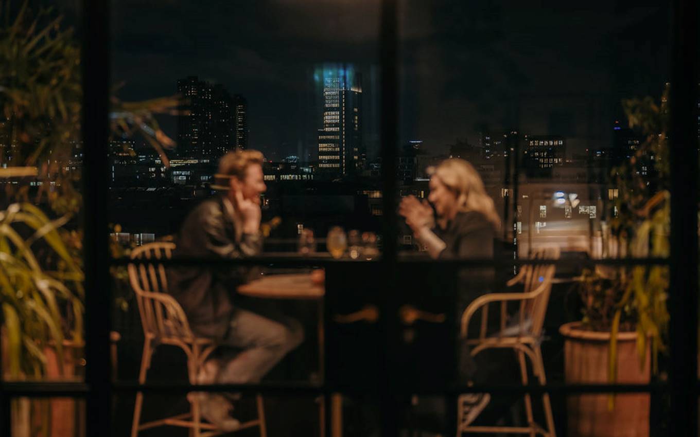 A couple seated at a rooftop restaurant in London, softly lit against a blurred city skyline at night.