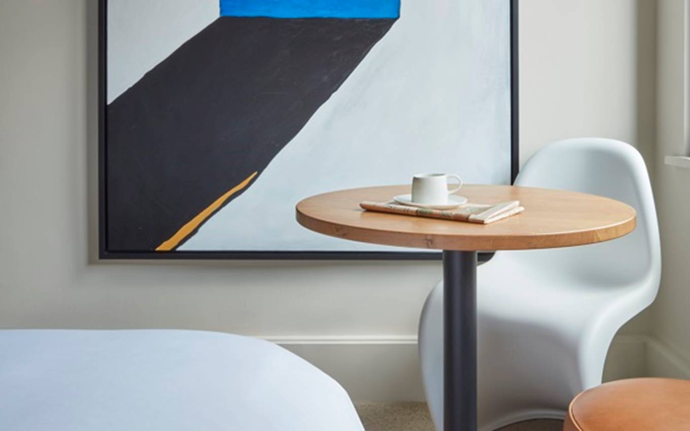 Modern art piece with bold blue and black shapes, hung beside a small wooden table and a white chair at a London hotel room.