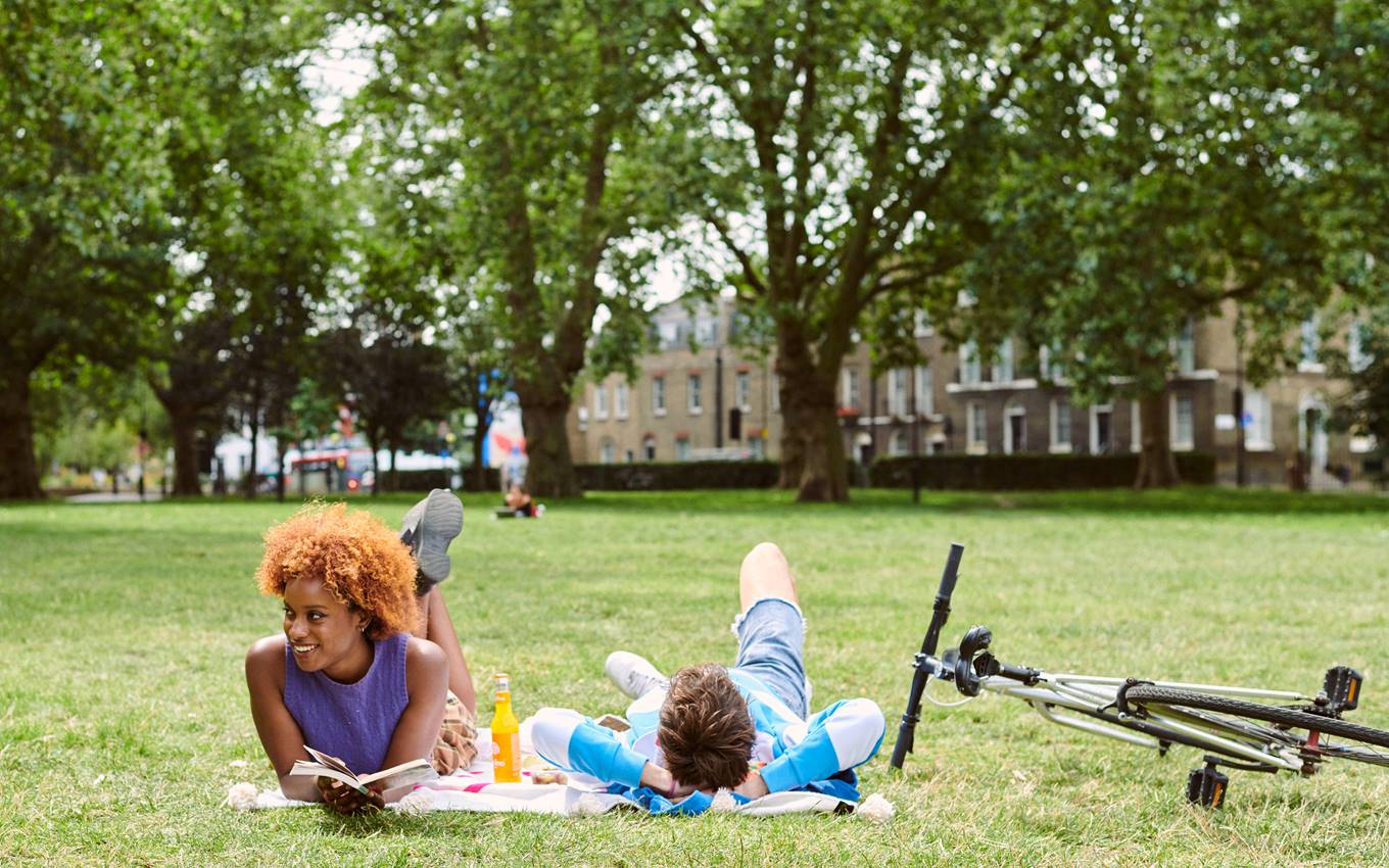 Two friends relaxing on a blanket in a London park, reading and chatting, with a bicycle nearby and trees surrounding them.