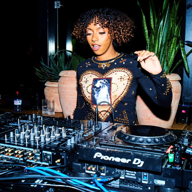 DJ performing at a Pioneer DJ setup, wearing a black top with gold accents and a heart design, plants in the background.