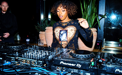 DJ performing at a Pioneer DJ setup, wearing a black top with gold accents and a heart design, plants in the background.
